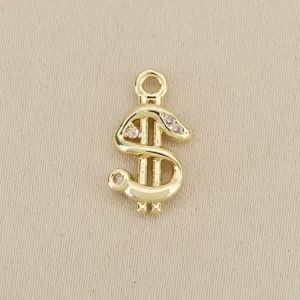 Close-up of a gold dollar-sign pendant with clear stones and a loop, laid on beige fabric