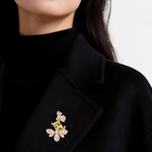 Spilla color oro rosa con gemma gialla rettangolare e dettagli bianchi, indossata su un cappotto nero
