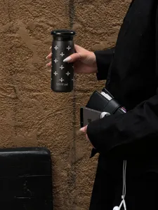 Una mano tiene una tazza termica nera con rilievo a stelle davanti a un muro di mattoni ruvido