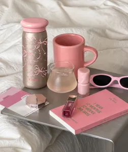 Bicchiere termico dorato con fiocchi rosa, disposto tra cosmetici e accessori rosa su un tavolo