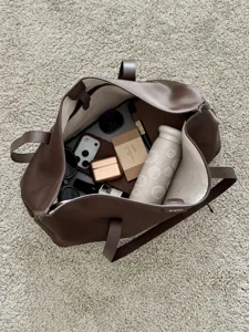 Vista dall'alto di una borsa tote marrone aperta con bottiglia termica color crema e accessori