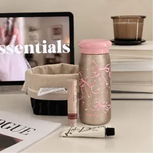 Scena su un tavolo con tazza termica color champagne, coperchio rosa e motivi di fiocchi e cuori