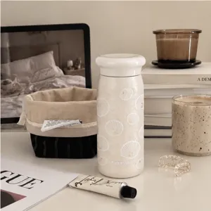 Tazza crema con conchiglie bianche e coperchio effetto ceramica, tra libri e decorazioni