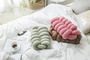 Three knot pillows in green, pink, and brown shown on a cozy white bed setting
