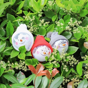 Three snowman Christmas tree ornaments with knit hats set in green garden bushes