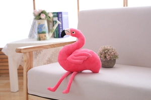 Pink flamingo plush toy with long legs sitting on a cozy grey fabric sofa in a home setting