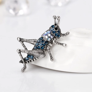 Vintage silver grasshopper brooch with blue rhinestones on a white ceramic display