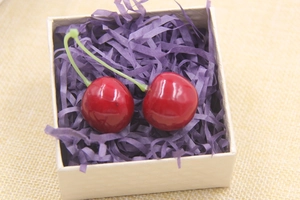 Detail showing two glossy artificial red cherries inside a gift box with purple shredded paper