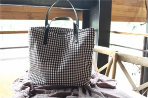 Detail of plaid canvas tote bag with black leather straps and metal rivets on a wooden table