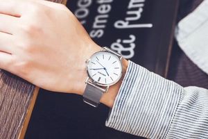 Close-up of a minimalist silver mesh strap watch on a wrist with a white dial and blue hands