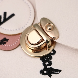 Polished gold metal flip lock hardware on minimalist cream leather bag with embroidery