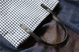 Detail of blue and white checkered canvas tote bag with black leather straps and gold rivets