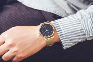 Close-up of a gold Milanese mesh strap watch with a black dial worn on a striped shirt