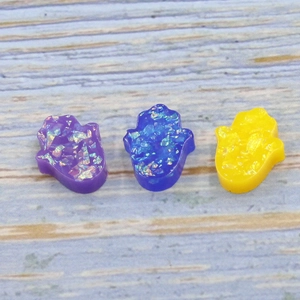 Three Hamsa hand beads in purple, blue, and yellow resin with glitter on blue wood background