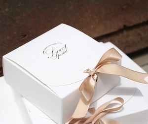 Close-up of a white square gift box with a champagne gold ribbon bow and gold lettering