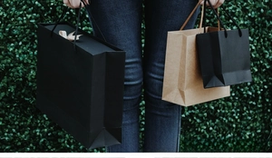 Person holding black and kraft paper gift bags with rope handles against a green hedge