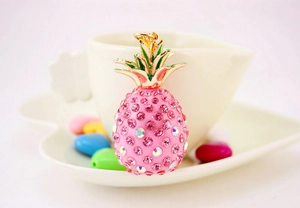 Close-up of pink rhinestone pineapple keychain with golden leaves near a white coffee cup