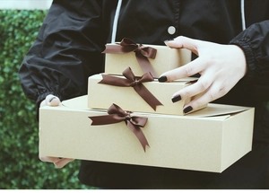 Three-piece set of brown kraft gift boxes with dark brown satin ribbon bows in a natural hand-held view
