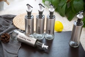 Set of glass and stainless steel oil dispensers on wood, featuring clear vinegar and sauce labels