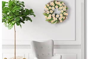White daisy wreath with green leaves on brown rattan base hanging on a minimalist wall