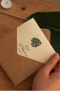 Detail showing hand holding beige greeting card with monstera leaf and kraft envelope