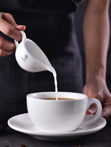 Close-up of white ceramic milk pitcher pouring liquid into a cup on dark background