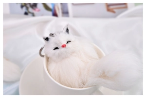 Close-up of a white plush fox keychain resting inside a ceramic mug with soft lighting