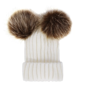 Close-up of white knitted baby hat with two large brown faux fur pom-poms on top
