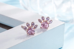 Macro shot of rose gold paw print studs with pink heart-shaped crystals on white stand