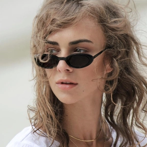 Minimalist black oval sunglasses with dark lenses worn by a model in outdoor setting