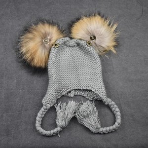 Gray baby knit trapper hat with two large raccoon fur pom-poms and braided ear flaps