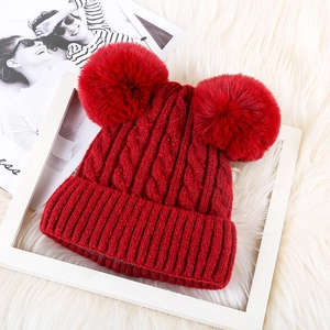 Close-up of a red knitted winter hat featuring fluffy double pom-poms on a white faux fur rug