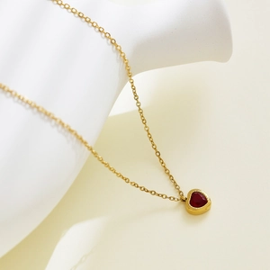 Detail shot of a gold heart necklace with a vibrant red zircon stone on a white ceramic stand