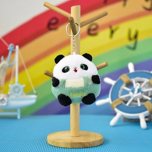 Close-up of a cute panda plush keychain wearing green Lucky overalls on a wooden stand.