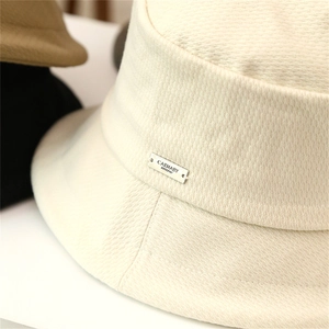 Close-up of beige textured fisherman hat with signature silver metal logo plaque detail