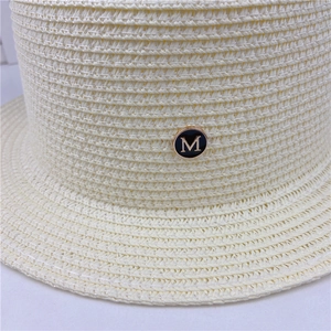 Detail of beige straw hat featuring a black circular metal emblem with gold letter M
