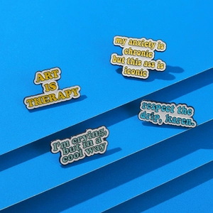 Detailed close-up of Art is Therapy and humorous enamel pins on a blue tiered background