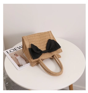 Close-up of a woven straw handbag featuring a large black ribbon bow on a white table