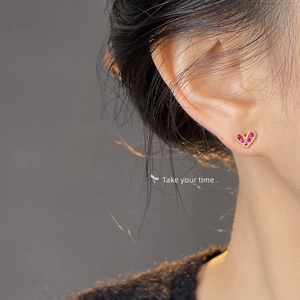Close-up of heart-shaped gold studs with red zircon and beaded texture on model ear