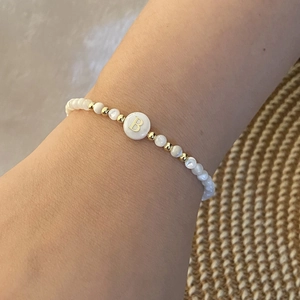 Detail of beaded bracelet with white shell beads and golden letter B charm on straw background