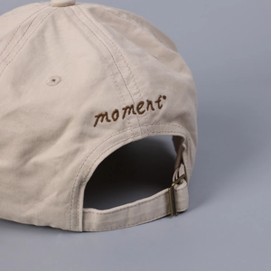 Detail of beige cotton baseball cap featuring moment embroidery and adjustable bronze buckle