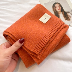 Close-up of soft orange knitted scarf with emo logo tag held by hand on white bedsheet