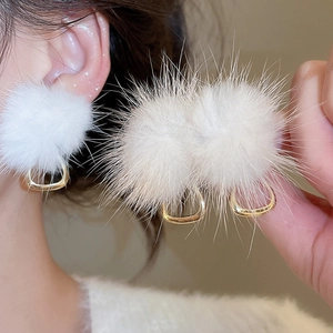 Close-up of white plush ball earrings with gold trapezoid metal accents worn by a model