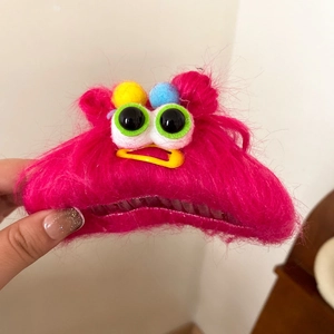 Pink plush monster hair claw clip with big eyes and colorful pom-poms being held in hand