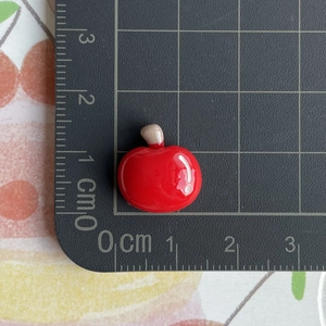 Macro shot of a red resin apple charm on a scale mat showing its 1.5cm width and 1.3cm height