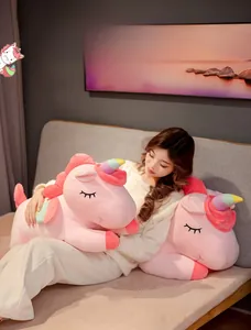 Pink unicorn plush pillow with rainbow horn and wings shown held by a person in bed