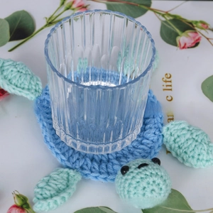 Detail of a handmade crochet turtle coaster under a ribbed glass on a floral background