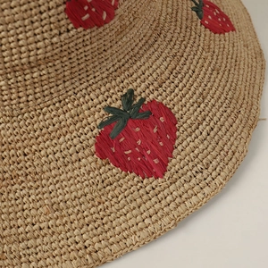 Close-up detail of handcrafted red strawberry embroidery on a natural raffia straw sun hat.