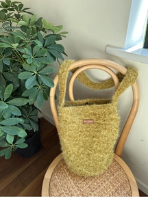 Detail einer grünen Plüsch-Schultertasche mit Lederlogo auf einem Holzstuhl