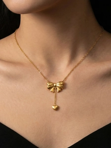Close-up of model wearing gold bow and heart pendant necklace against black background